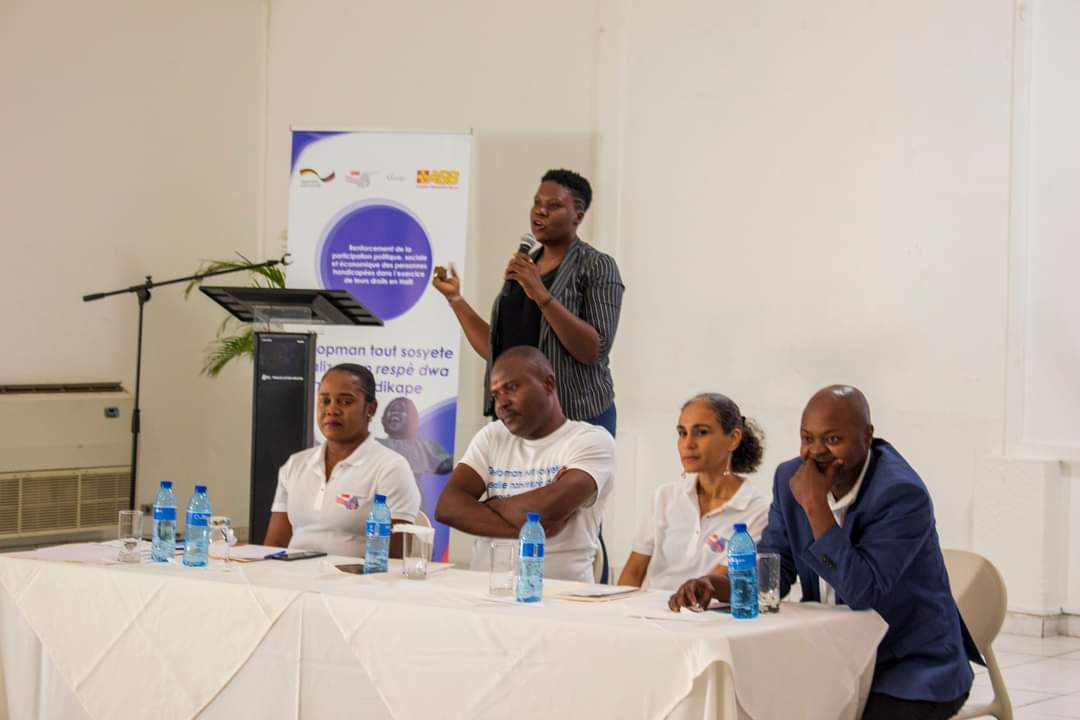 Photo d'une salle de cours en Haïti où des adultes sont assis à des pupitres d'écoliers et semblent écouter un interlocuteur hors cadre